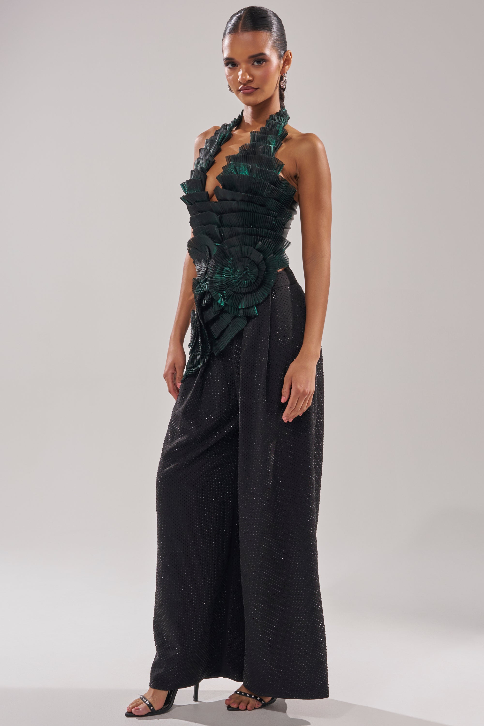 A woman poses in the DREAMERS HALTER BLOUSE, a sculptural black organza top with emerald green pleated rosettes and wide-leg pants. She completes the look with strappy heels and her hair pulled back.