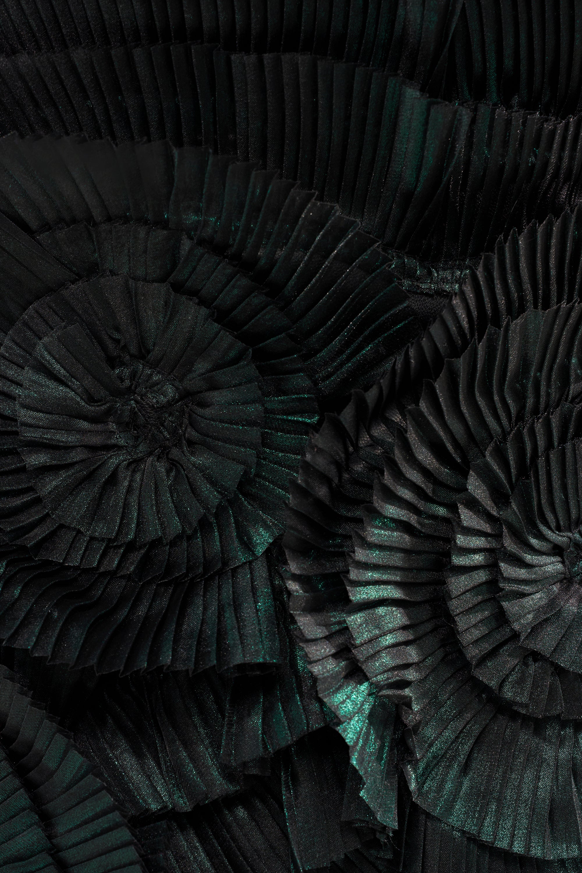Close-up of pleated, dark green and black fabric arranged in spirals, echoing the textured, abstract design of the DREAMERS HALTER BLOUSE, with subtle shimmering highlights enhancing its striking look.