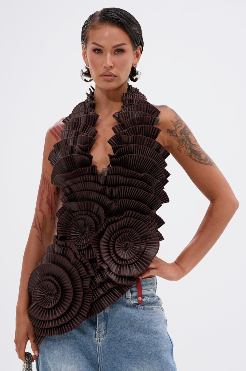 A woman exudes confidence in the DREAMERS BLOUSE, a sleeveless faux leather top with bold pleated rosettes, styled with light blue jeans. She sports slicked-back hair and displays tattoos on her upper arm.