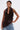 A woman exudes confidence in the DREAMERS BLOUSE, a sleeveless faux leather top with bold pleated rosettes, styled with light blue jeans. She sports slicked-back hair and displays tattoos on her upper arm.