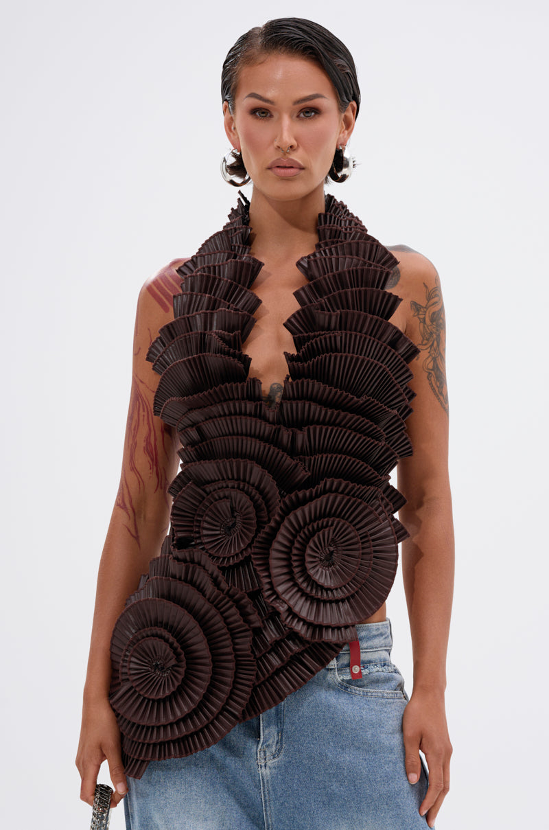 A person with short, slicked-back hair wears the DREAMERS BLOUSE—featuring large, dark brown pleated rosettes and a halter neckline—with loose blue jeans. Tattoos are visible on their arms as they pose against a plain white background.