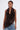 A person with short, slicked-back hair wears the DREAMERS BLOUSE—featuring large, dark brown pleated rosettes and a halter neckline—with loose blue jeans. Tattoos are visible on their arms as they pose against a plain white background.