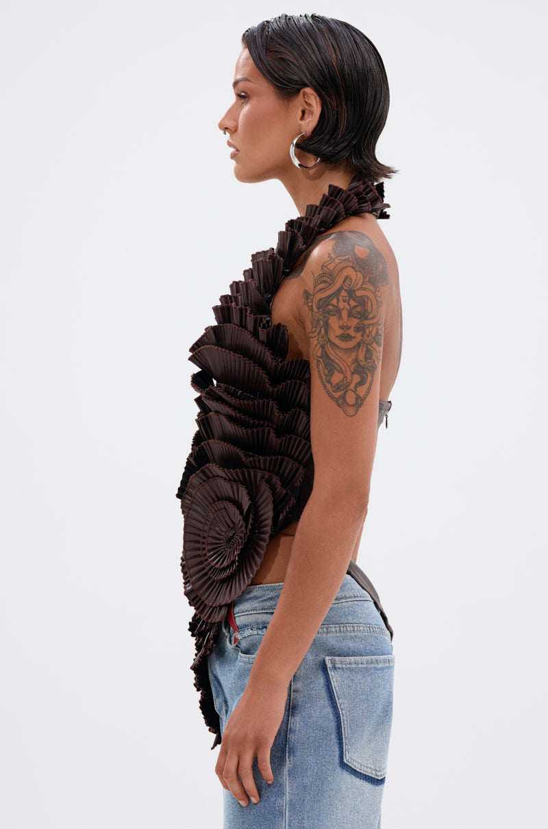 A woman in profile wears the DREAMERS BLOUSE, a sculptural faux leather top with pleated rosettes, paired with light blue jeans. She has short, slicked-back hair, hoop earrings, and a large upper arm tattoo against a white background.