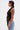 A woman in profile wears the DREAMERS BLOUSE, a sculptural faux leather top with pleated rosettes, paired with light blue jeans. She has short, slicked-back hair, hoop earrings, and a large upper arm tattoo against a white background.
