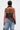 Seen from behind, a person with short, slicked-back hair wears the DREAMERS BLOUSE—a brown faux leather top with an asymmetrical halter neckline—paired with loose blue jeans. Their tattooed back and arms are streaked with red paint as they hold a silver chain bag.