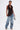 A person stands confidently in the DREAMERS BLOUSE—a faux leather top with pleated rosettes and a halter neckline—paired with loose blue jeans, a metallic purse, statement earrings, slicked-back hair, and heeled sandals.