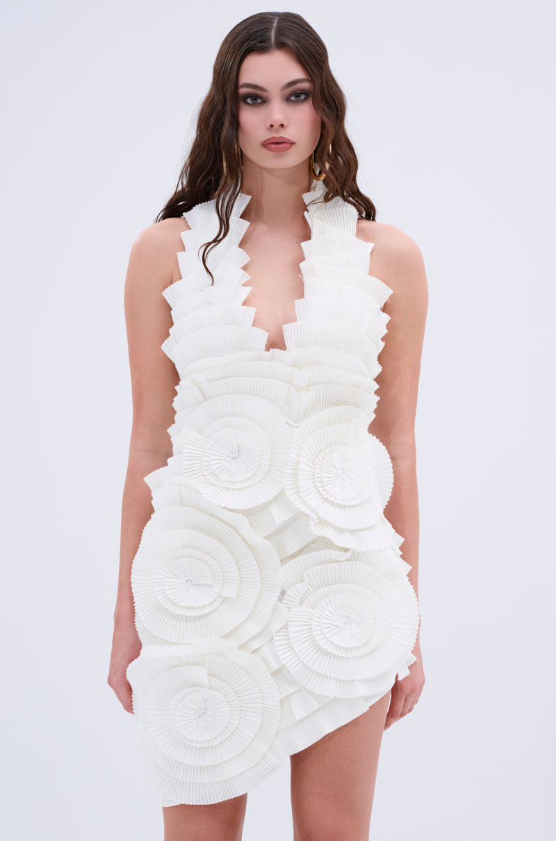 A woman with long wavy hair models the ONE OF ONE BANDAGE MINI DRESS IN WHITE, a short, sleeveless mini with sculptural circular ruffles, exuding avant-garde elegance against a plain light background.