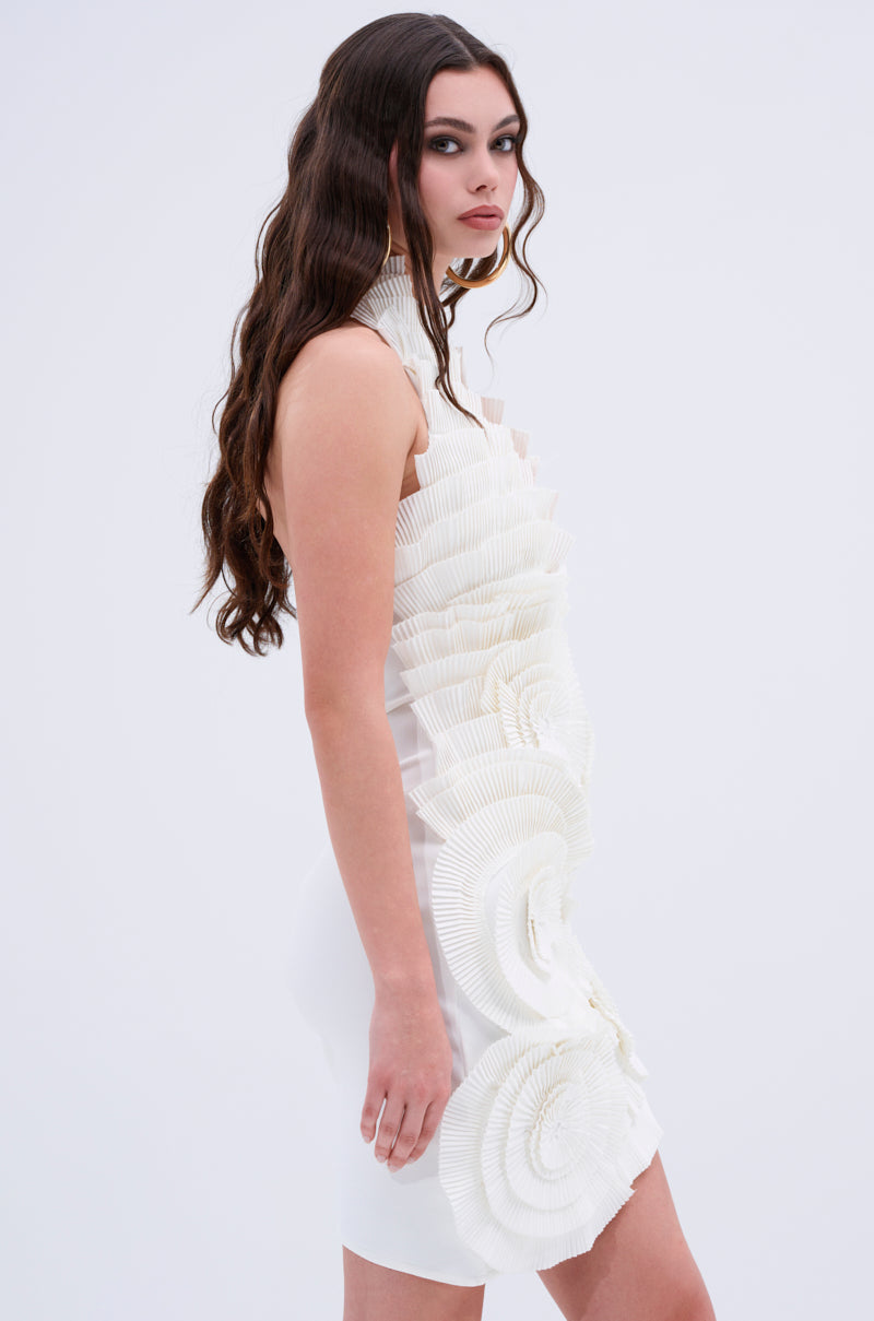 A woman with long wavy brown hair wears the ONE OF ONE BANDAGE MINI DRESS IN WHITE, featuring intricate, layered pleated details. She exudes sculptural elegance against a plain light background, glancing slightly over her shoulder.