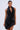 A woman stands against a plain, light background wearing the ONE OF ONE MINI DRESS IN PU, a unique sleeveless black mini dress with large pleated rosettes. Her hair is pulled back and she accessorizes with geometric earrings.