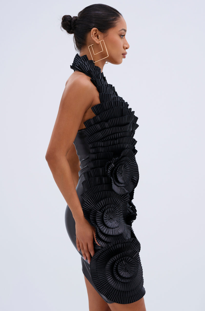 A woman poses in the ONE OF ONE MINI DRESS IN PU, a fitted, sleeveless black faux leather dress with bold pleated spiral accents; she wears her hair in a bun and accessorizes with geometric gold earrings.