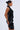 A woman poses in the ONE OF ONE MINI DRESS IN PU, a fitted, sleeveless black faux leather dress with bold pleated spiral accents; she wears her hair in a bun and accessorizes with geometric gold earrings.