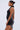 A woman in the ONE OF ONE MINI DRESS IN PU stands in profile. The black faux leather dress features dramatic pleated ruffle details. She wears her hair in a neat bun and large square earrings, set against a plain light background.