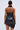 A woman with her hair in a neat bun is seen from the back wearing the ONE OF ONE MINI DRESS IN PU, a shiny black faux leather dress with an asymmetrical hem and textured details, set against a plain light background.