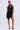 A woman stands confidently against a plain light background wearing the ONE OF ONE MINI DRESS IN PU, a sleeveless black mini dress with asymmetrical design, bold circular textures, pleated rosettes, and paired with black high-heeled sandals.
