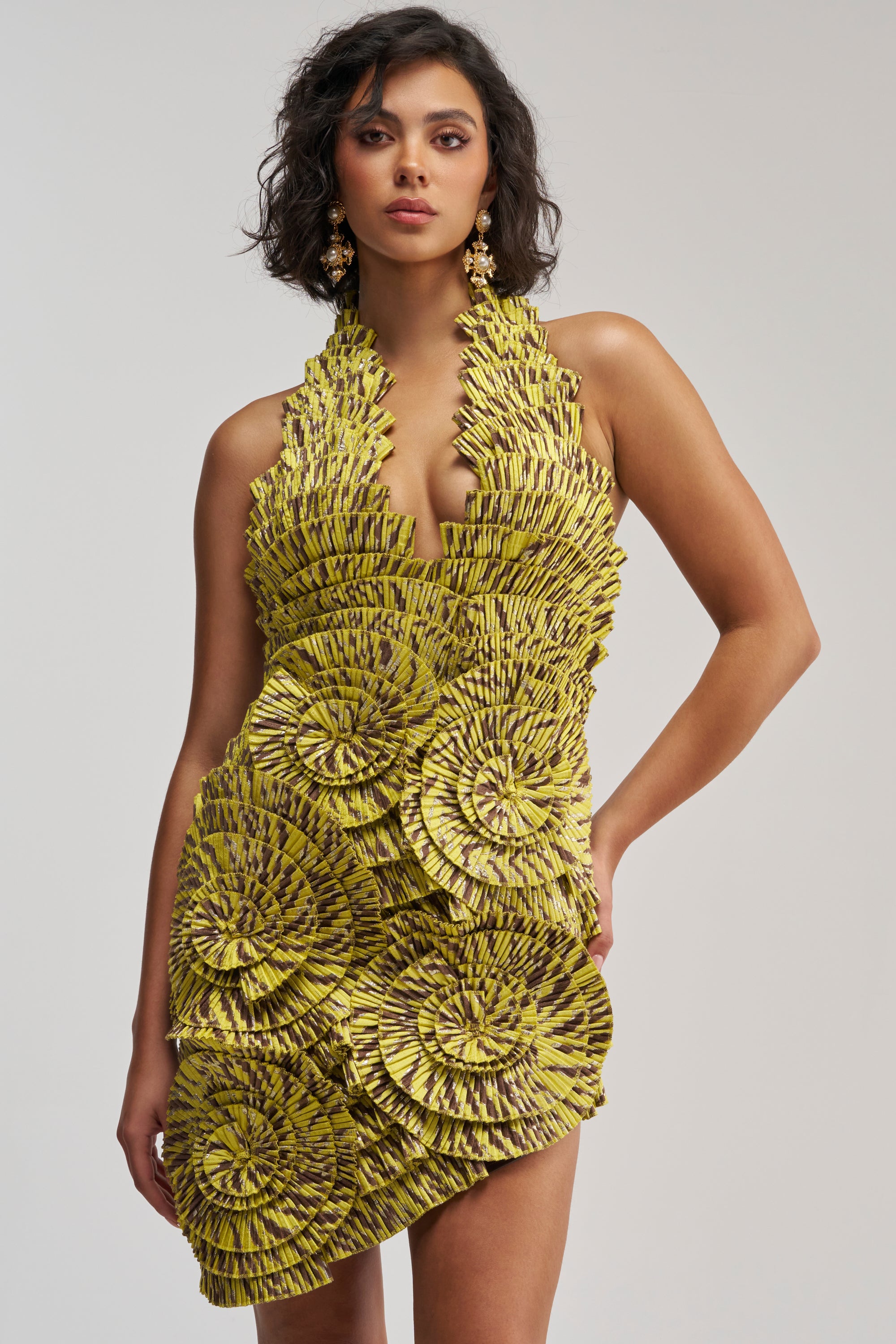 A woman stands against a plain light background wearing the SHOWING OFF NOVELTY MINI DRESS IN YELLOW MULTI—a sleeveless, geometric-textured mini dress featuring layered circular patterns, a deep V neckline, and an asymmetrical hem for a bold cocktail party look.