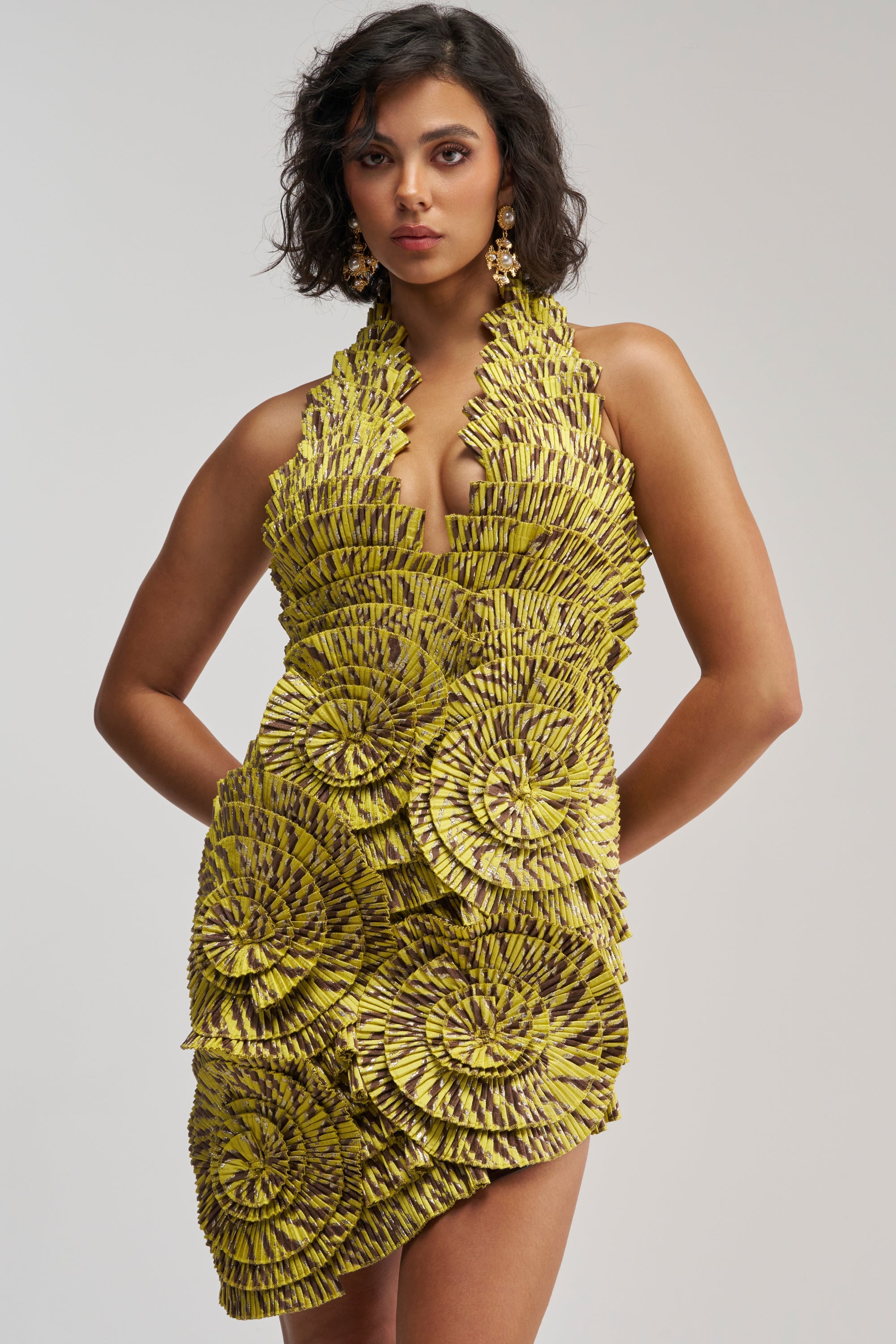 A woman stands with hands on hips against a plain white background, wearing the SHOWING OFF NOVELTY MINI DRESS IN YELLOW MULTI—a sculpted, sleeveless cocktail party mini dress featuring bold yellow and black colors with large circular patterns and textured detailing.