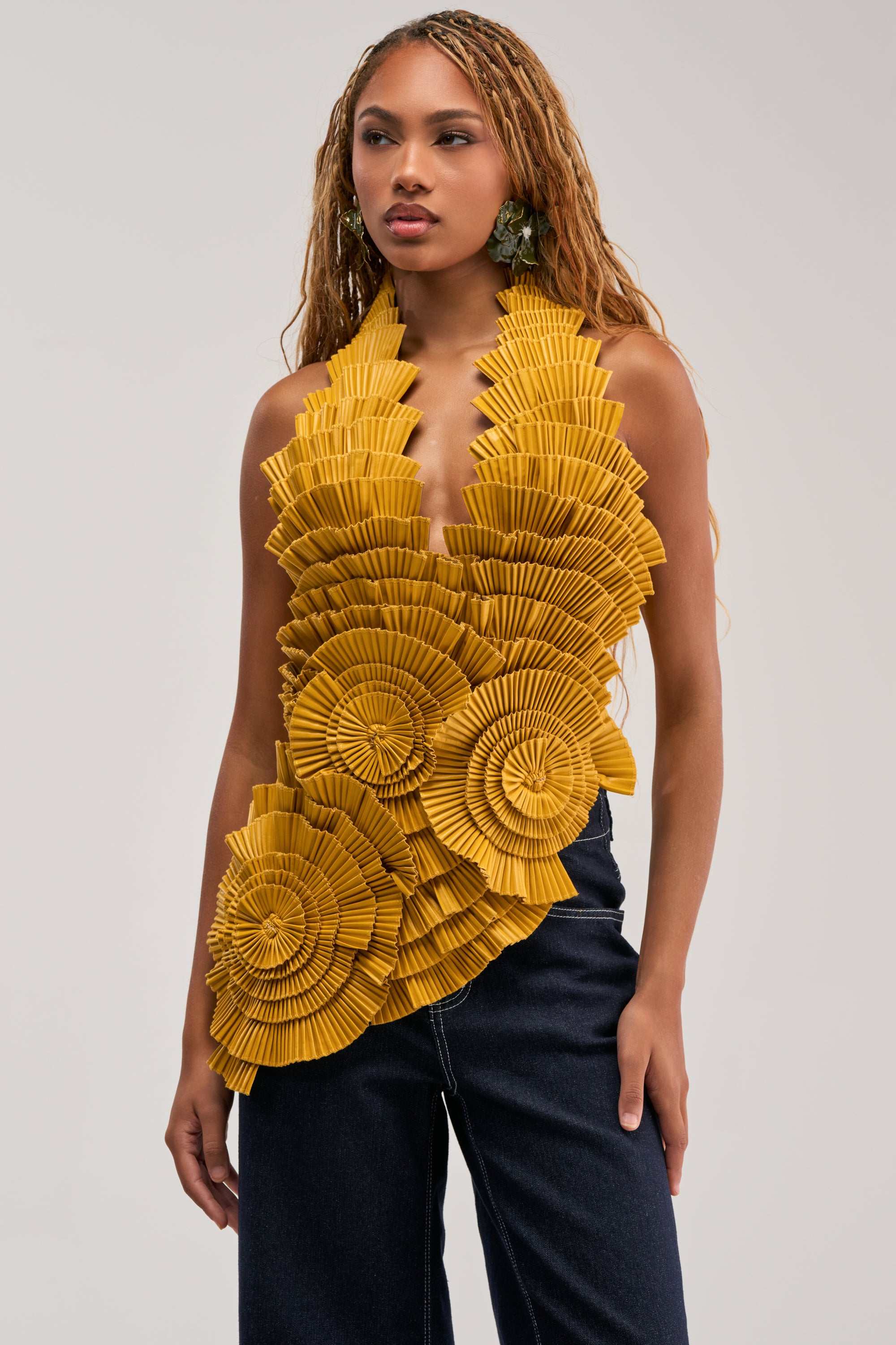 A woman wears the MOLTEN PETAL PLEATED TOP—a sleeveless, yellow sculptural blouse featuring large, pleated, fan-like ruffles—paired effortlessly with dark jeans as she stands against a plain light gray background.