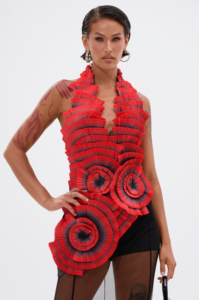 A person poses confidently in the CRAZY IN LOVE BLOUSE, a striking pleated rosette top with black accents and layered spiral designs. They wear a sheer black skirt, have short slicked-back hair, visible tattoos, and stand before a plain white background.