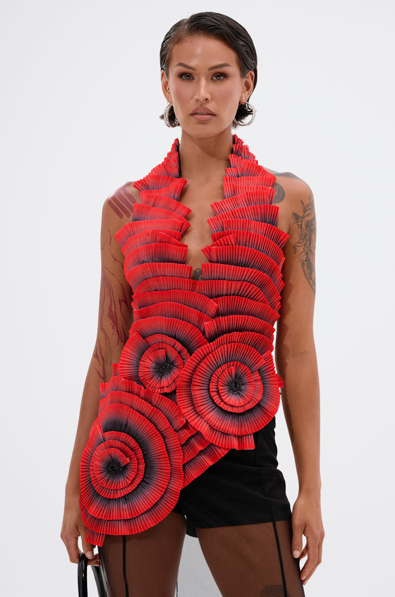 A person with wet-look hair wears the bright red CRAZY IN LOVE BLOUSE with sculptural pleats, circular patterns, and black accents, paired with black shorts, standing against a plain white background.