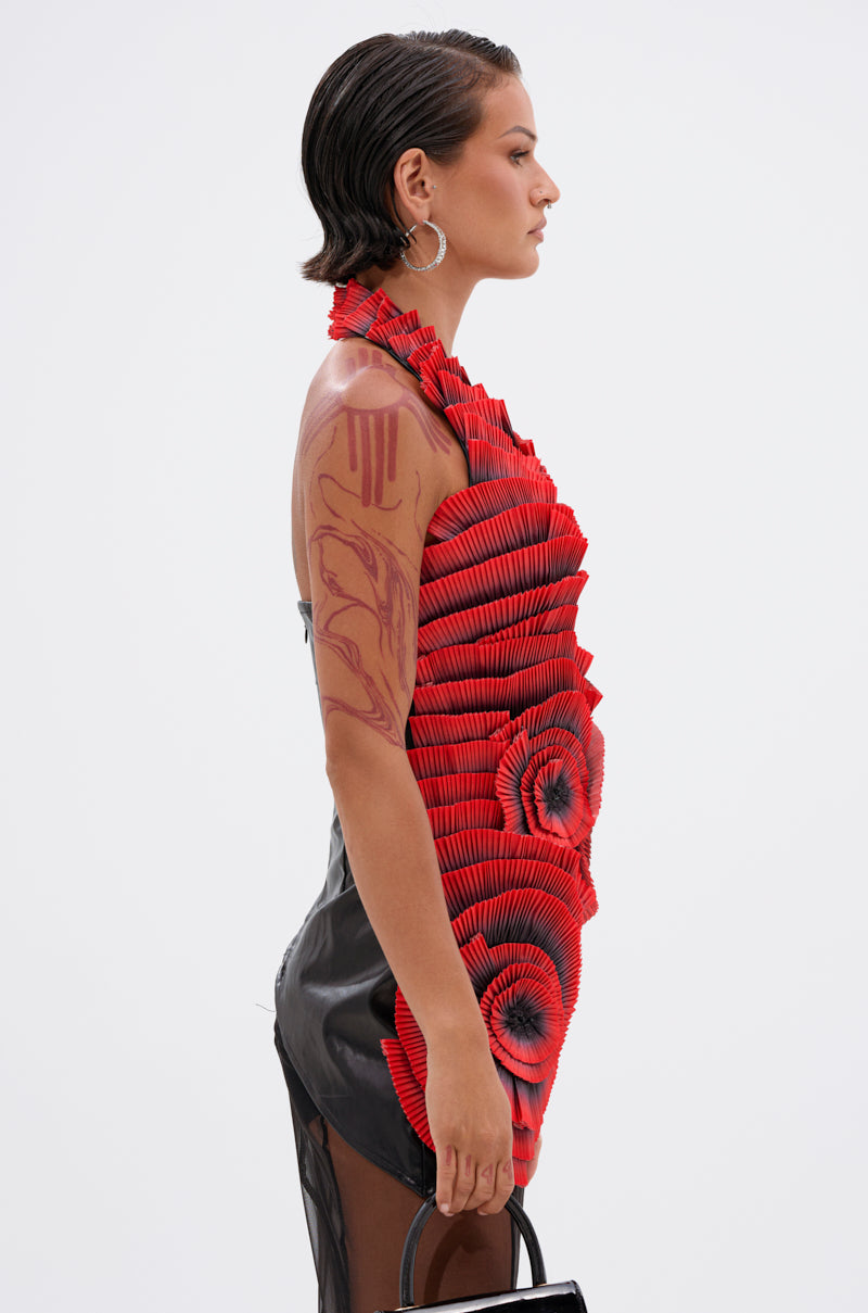 A woman in profile showcases the CRAZY IN LOVE BLOUSE, a sculptural pleated rosette top with red and black circular patterns. She sports slicked-back hair, large hoop earrings, visible arm tattoos, and holds a black handbag against a plain white background.