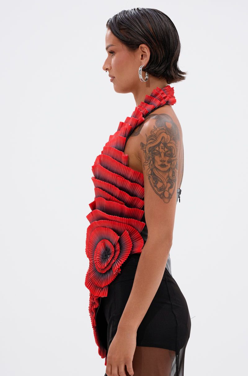 A woman with short dark hair, hoop earrings, and an upper arm tattoo stands in profile wearing the CRAZY IN LOVE BLOUSE in red and black with a sheer black skirt, set against a plain white background.