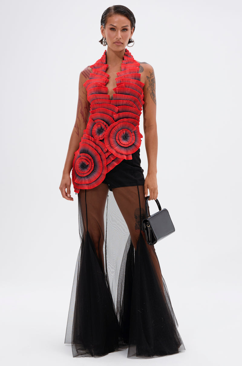 A person wears the CRAZY IN LOVE BLOUSE, featuring bold red pleated rosettes, paired with a black sheer skirt over shorts and holding a small black handbag, against a plain white background.