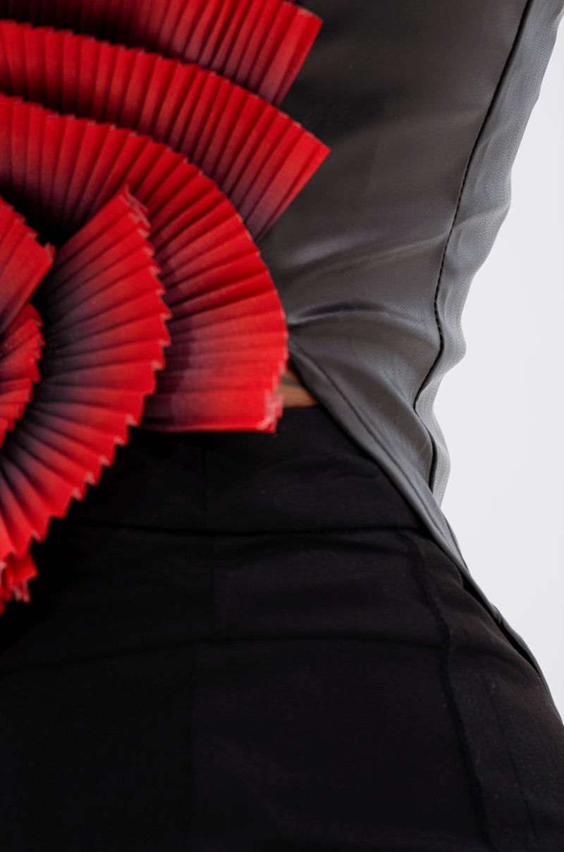 A close-up of a person wearing the CRAZY IN LOVE BLOUSE, a fitted black top with a plunging halter neckline and bold sculptural red pleated detail on one side for a striking, modern look.