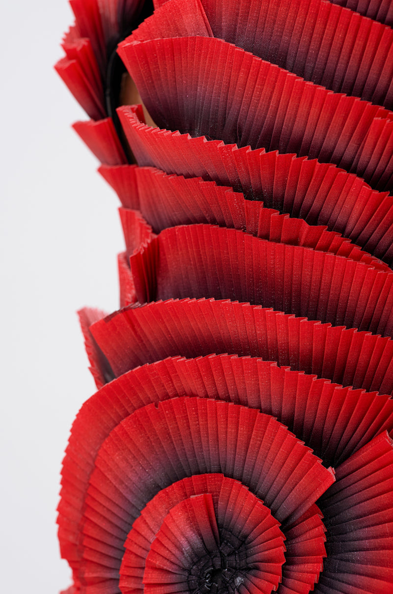 Close-up of the CRAZY IN LOVE BLOUSE with overlapping, fan-shaped red ombre pleated fabric layers arranged in a spiral pattern against a light background.