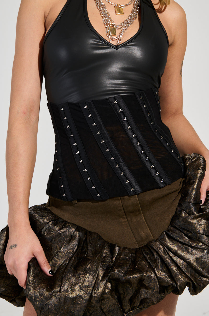 A person showcases a black leather top adorned with studded accents, complemented by layered necklaces and a skirt featuring textured ruffles. Their outfit is perfectly accessorized with the CINCH ME UP STRETCH BELT, which includes faux leather trim. An elegant tattoo is subtly visible on their left wrist. 1