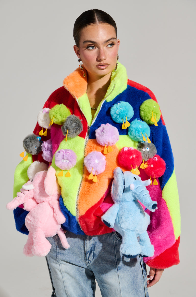 A person wearing the HERE FOR A GOOD TIME TRINKET BOMBER, a vibrant multicolored faux fur jacket adorned with plush toys featuring small round animals and two large elephants, stands against a plain background. Their hair is tied back, and they are wearing jeans.