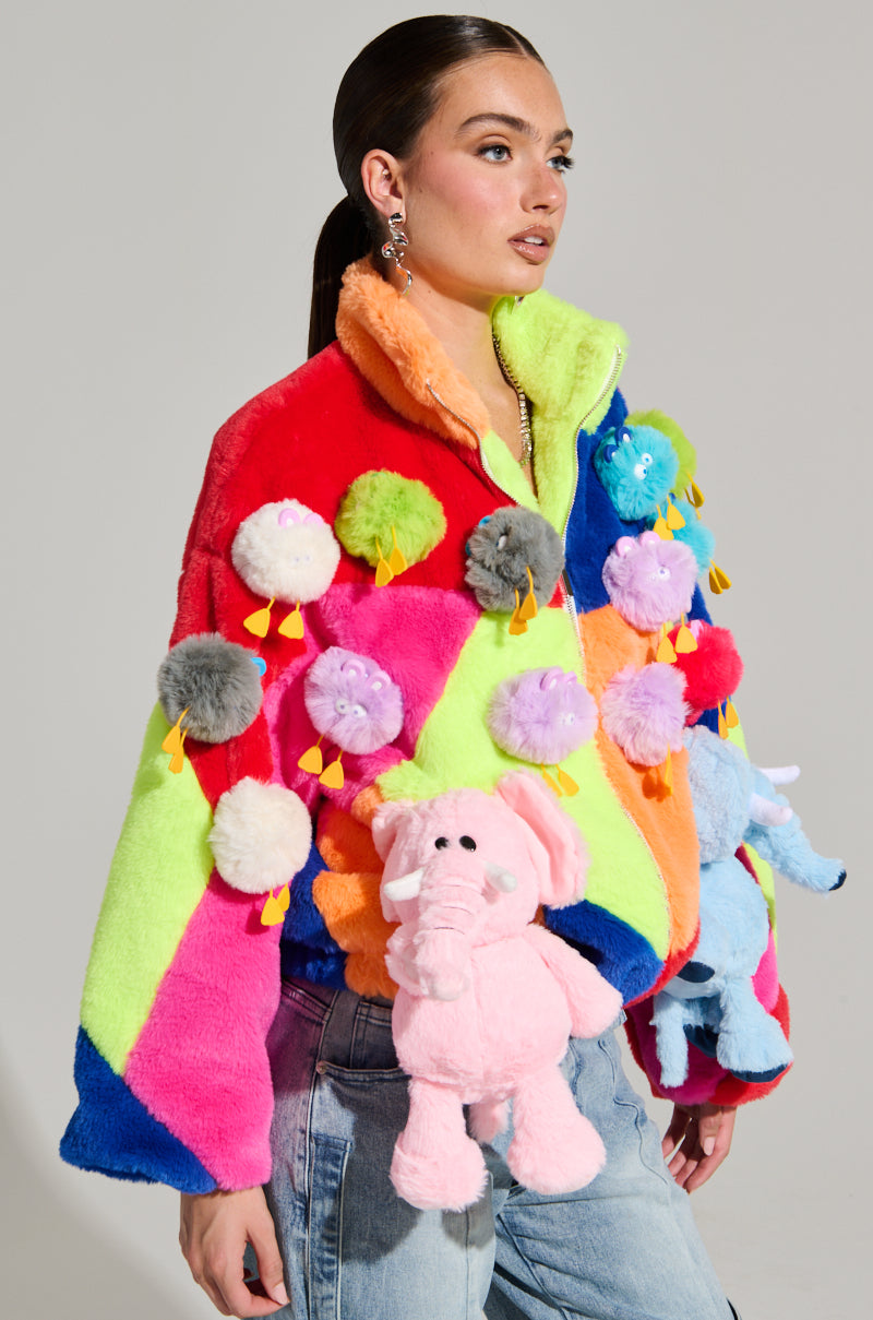 A model displays the HERE FOR A GOOD TIME TRINKET BOMBER, a vibrant jacket with colorblock paneling, embellished with multicolored pom-poms and plush animal figures like a pink elephant. Her slicked-back hair and jeans perfectly highlight the faux fur bomber's bold rainbow tones such as red, green, and blue.