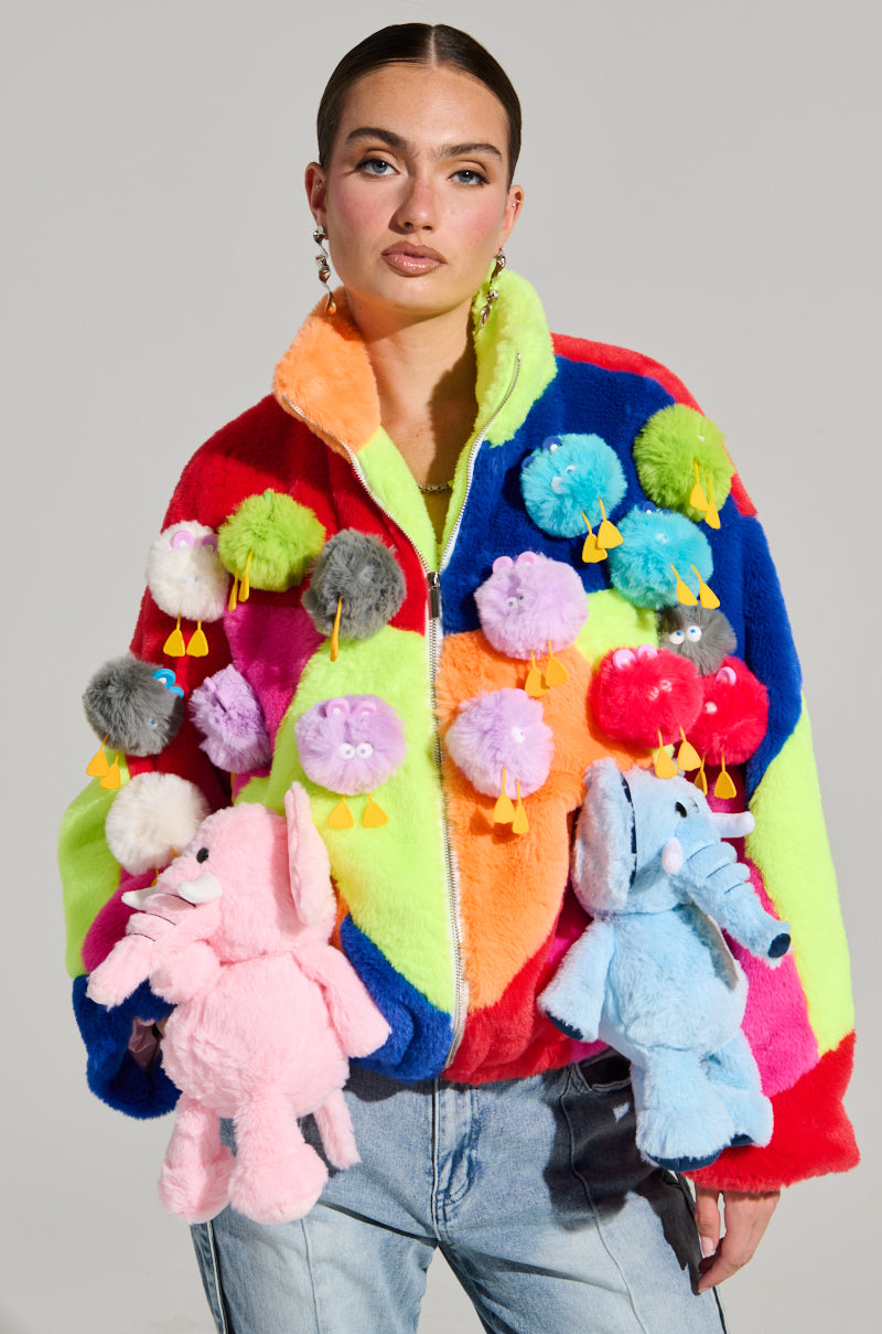 Wearing the HERE FOR A GOOD TIME TRINKET BOMBER, adorned with plush animal keychains like elephants and various round creatures, the person showcases vibrant colorblock paneling in red, blue, green, and orange. The jacket is paired with light blue jeans.
