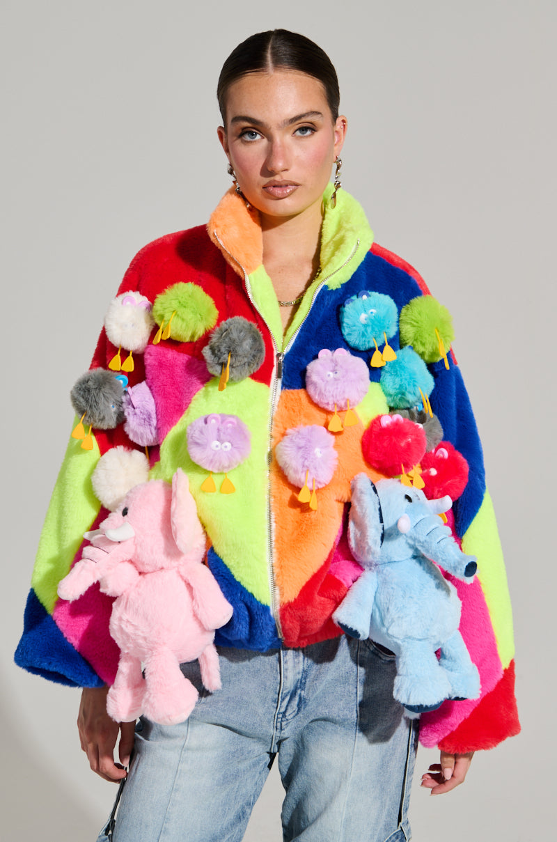 Against a plain background, a person stands out in the HERE FOR A GOOD TIME TRINKET BOMBER, an eye-catching jacket adorned with plush animals and pom-poms. The vibrant colorblock paneling of the bomber bursting with bright hues such as pink, blue, green, and orange makes an ideal pairing with their light blue jeans.
