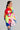 A person is wearing the HERE FOR A GOOD TIME TRINKET BOMBER, a jacket featuring vibrant color blocks and adorned with plush toys and pom-poms, showcasing dynamic shades of pink, red, yellow, green, and blue. Complementing their long cascading hair with hoop earrings and pairing it with jeans creates a playful vibe.