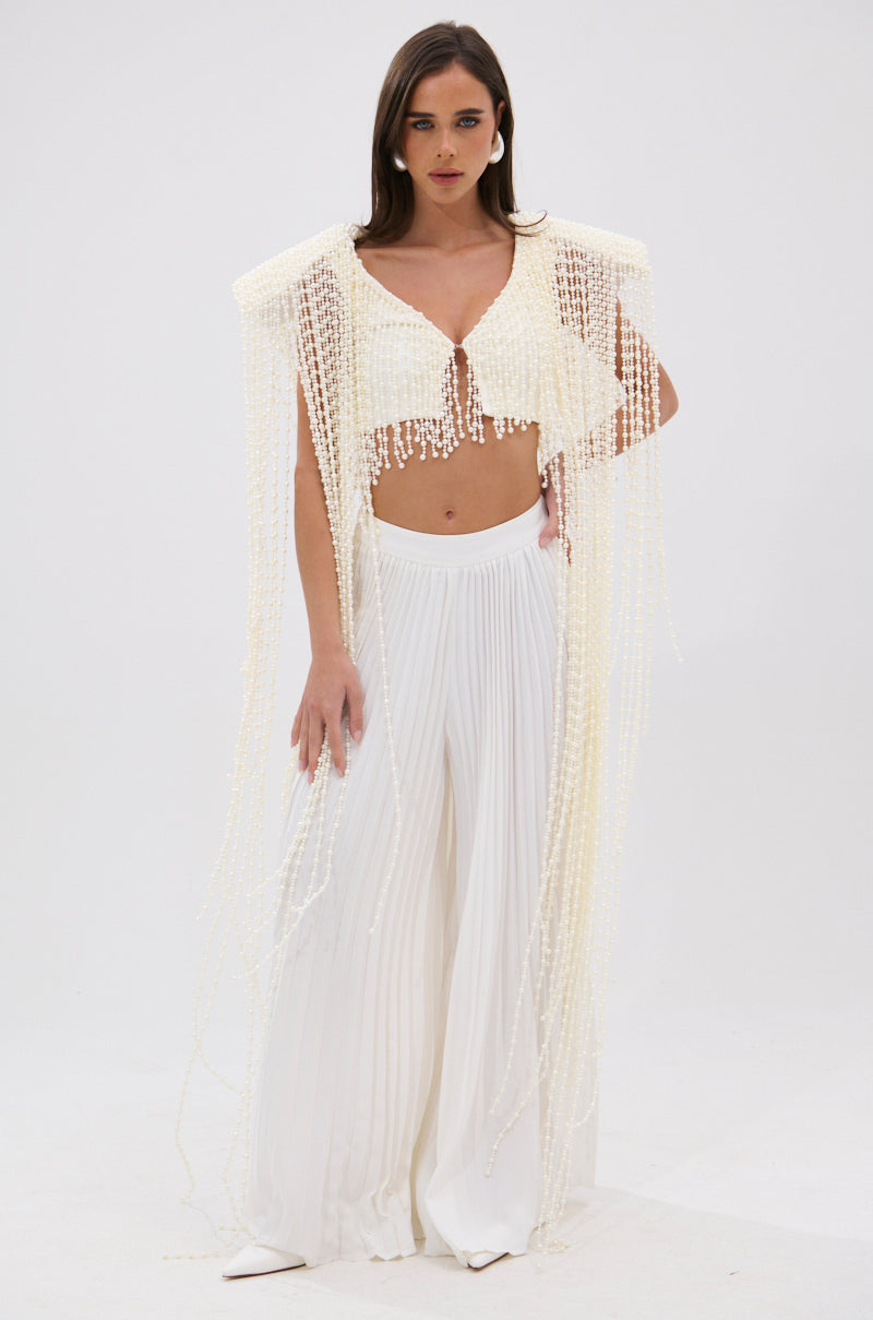 A woman with long brown hair and neutral makeup poses against a white background wearing the PEARL HEAVEN BEADED TOP, which features padded shoulders, paired with wide-legged, pleated white pants. Her hand rests by her side. 1