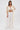 A woman with long brown hair and neutral makeup poses against a white background wearing the PEARL HEAVEN BEADED TOP, which features padded shoulders, paired with wide-legged, pleated white pants. Her hand rests by her side.