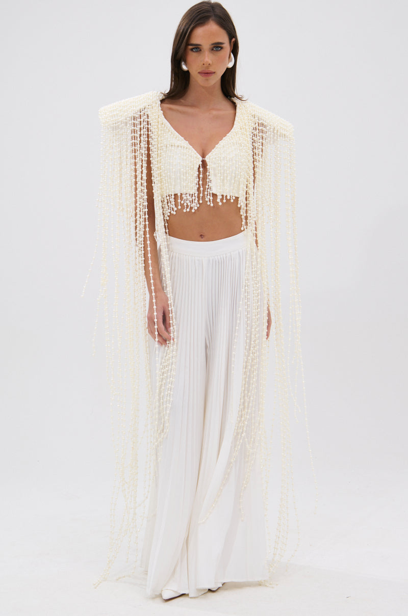 A woman with her hair down and a neutral expression models the PEARL HEAVEN BEADED TOP, featuring padded shoulders and lavish pearl beading, paired with wide-leg, pleated white pants against a white background. 2