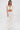 A woman with her hair down and a neutral expression models the PEARL HEAVEN BEADED TOP, featuring padded shoulders and lavish pearl beading, paired with wide-leg, pleated white pants against a white background.