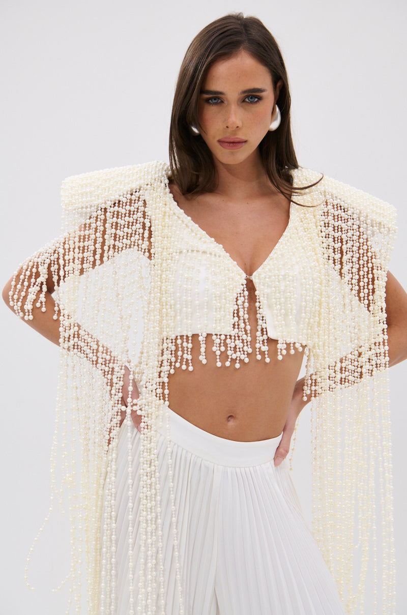 A woman with long brown hair models the PEARL HEAVEN BEADED TOP, featuring opulent pearl beads and padded shoulders, styled with matching statement earrings and high-waisted pleated white pants against a plain backdrop. 3