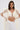 A woman with long brown hair models the PEARL HEAVEN BEADED TOP, featuring opulent pearl beads and padded shoulders, styled with matching statement earrings and high-waisted pleated white pants against a plain backdrop.