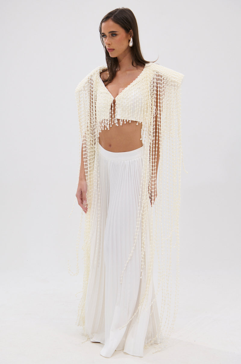 A woman poses elegantly against a plain white background in the PEARL HEAVEN BEADED TOP, featuring opulent pearl beads and padded shoulders, paired with high-waisted, wide-leg white pleated pants. 4