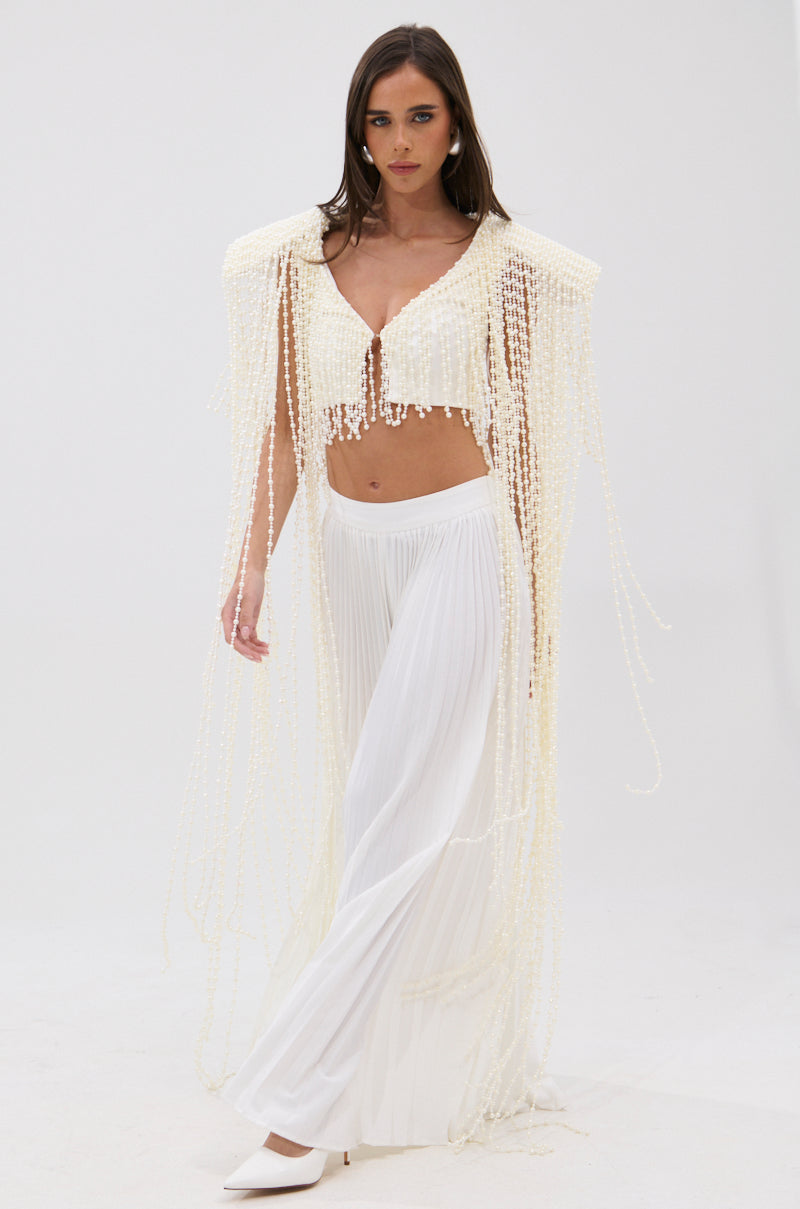 A woman wearing the PEARL HEAVEN BEADED TOP with padded shoulders and pearl embellishments, paired with wide-leg sheer pants and white pointed heels, stands against a plain white background. She has long brown hair. 0