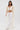 A woman wearing the PEARL HEAVEN BEADED TOP with padded shoulders and pearl embellishments, paired with wide-leg sheer pants and white pointed heels, stands against a plain white background. She has long brown hair.