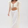 A woman wearing the PEARL HEAVEN BEADED TOP with padded shoulders and pearl embellishments, paired with wide-leg sheer pants and white pointed heels, stands against a plain white background. She has long brown hair.