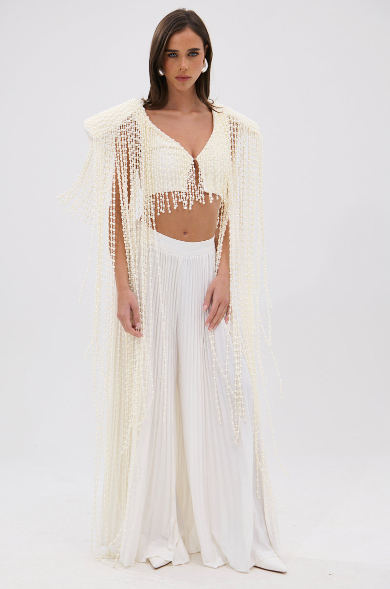 A woman stands against a white background wearing high-waisted wide-leg pants and the PEARL HEAVEN BEADED TOP, which features padded shoulders and a dramatic overlay of opulent pearl beads draping from her shoulders and arms. 5
