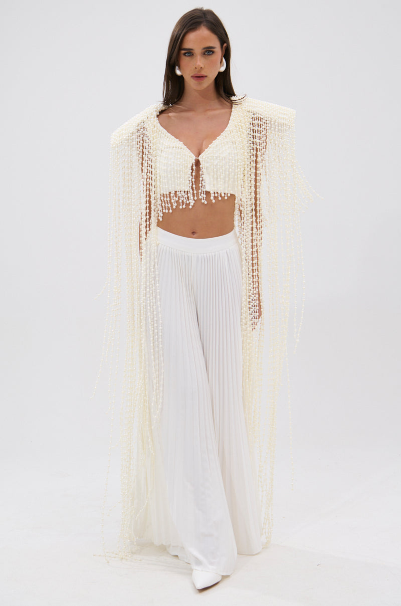 A woman with straight brown hair and a serious expression stands against a white background, wearing pleated wide-leg pants and the PEARL HEAVEN BEADED TOP, which has padded shoulders and dramatic cape-like pearl beading. 6