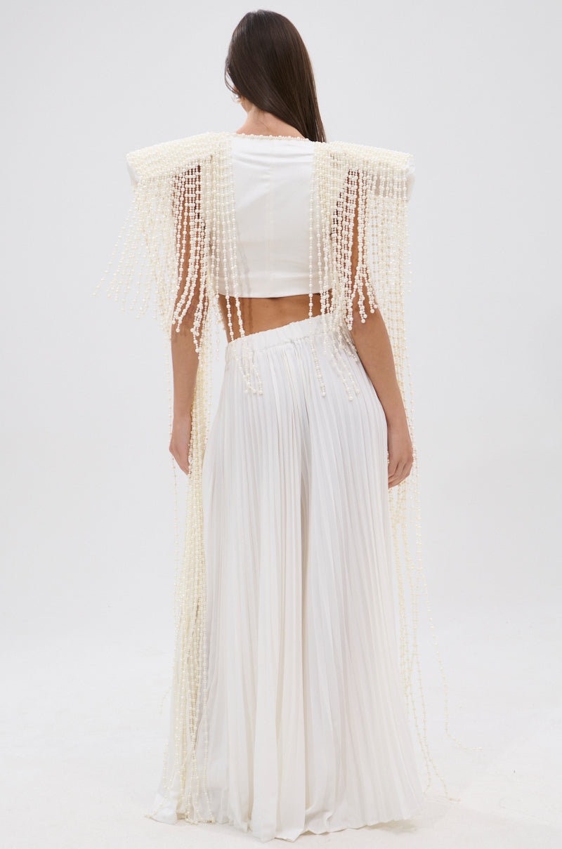 A woman stands facing away, wearing the PEARL HEAVEN BEADED TOP with padded shoulders and cascading pearl fringe, paired with a long pleated white skirt against a plain white background. 7