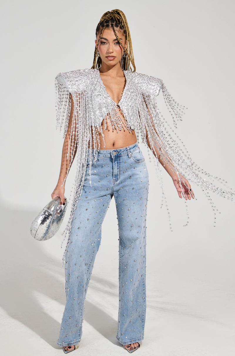 A person poses confidently in the IN MY OWN LANE SILVER BEADED TOP, complete with fringe detailing and beaded tassels, partnered with blue jeans embellished with silver studs for a perfect party look. They hold a matching silver clutch.
