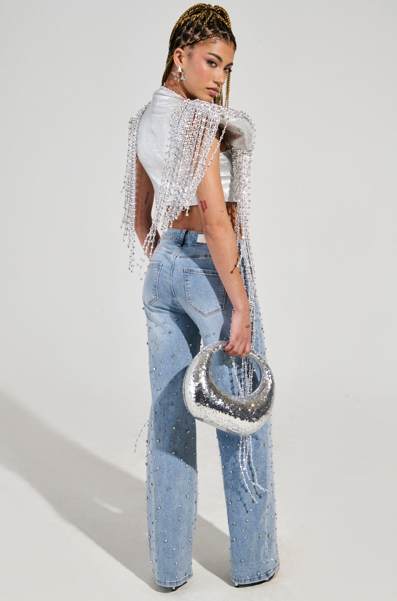 A woman with braided hair stands in profile, showcasing a white crop top with fringe details paired with light blue jeans featuring small embellishments. Her look is enhanced by the standout piece—a dazzling IN MY OWN LANE SILVER BEADED TOP—set against a neutral background.