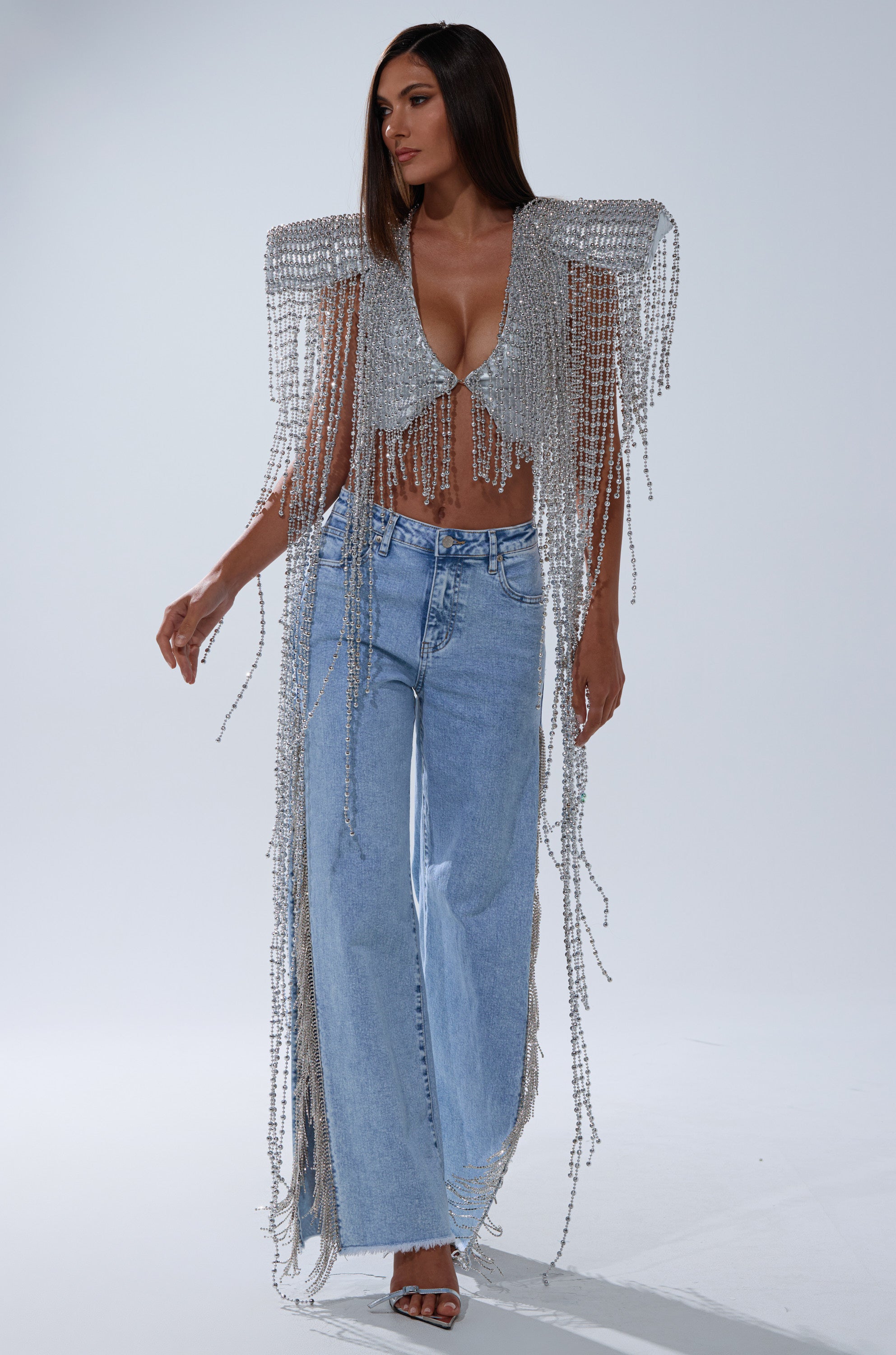 A woman stands against a plain light background wearing the IN MY OWN LANE SILVER BEADED TOP with dramatic shoulders and beaded fringe, paired with light blue wide-leg jeans and black open-toe heels.